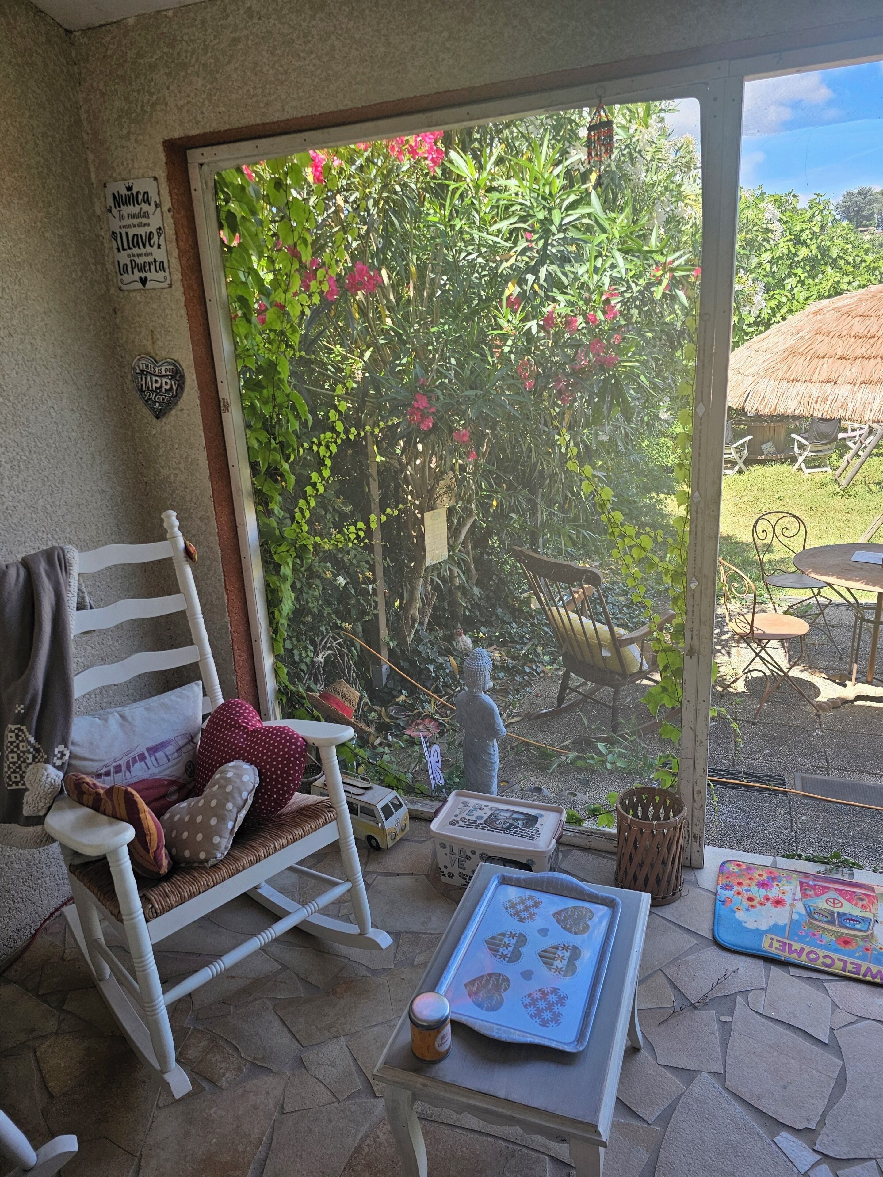 A cozy patio with a rocking chair, a small table, and a view of a garden through a glass door.