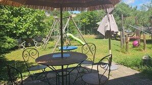 A thatched umbrella table with metal chairs, a swing set, and a playground in the background. - Cosy interlude at Anastasia's sunny home in the South of France (Bagnols-sur-Cèze)