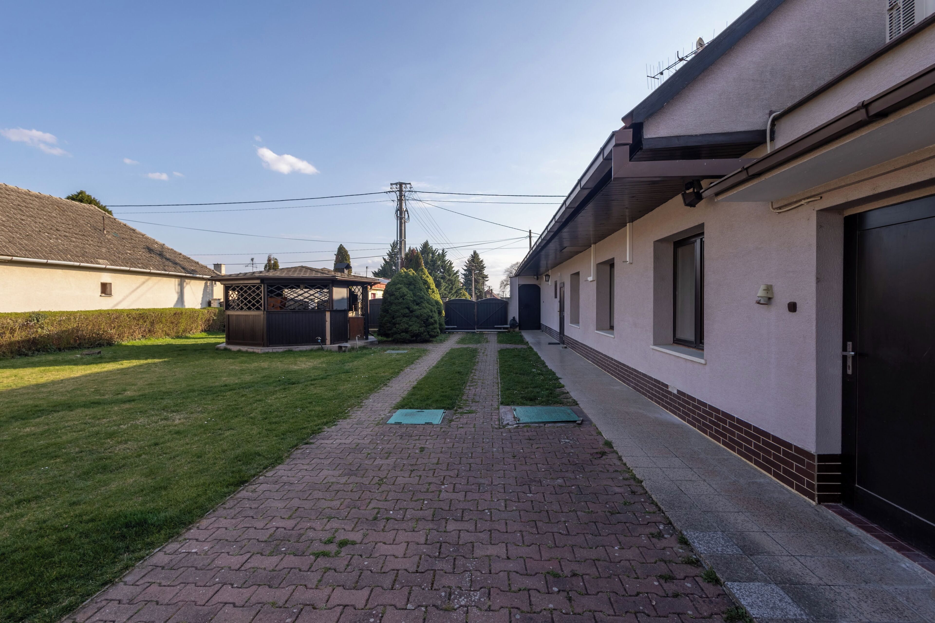 Un sendero pavimentado que conduce a un edificio con puerta negra, un pequeño jardín y cielo despejado.