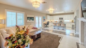 A spacious living area with a sofa, coffee table, and a floral arrangement. There is a kitchen with white cabinets and a bar area with stools. - Elegant 5BR Gem @Ontario w/ Grill | Near Airport (Ontario)