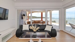 Ein modernes Wohnzimmer mit einer Couch, einem Couchtisch und einem Fernseher. Ein großes Fenster bietet Blick auf das Meer.
