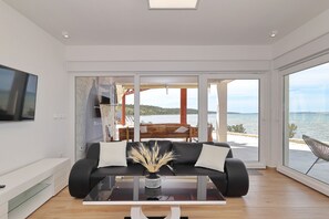 Ein modernes Wohnzimmer mit einer Couch, einem Couchtisch und einem Fernseher. Ein großes Fenster bietet Blick auf das Meer.