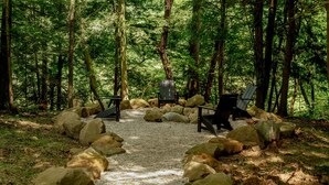 Uma área arborizada com um caminho de cascalho, cercada por pedras grandes, que leva a duas cadeiras pretas e uma mesa.