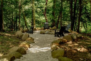 A forested area with a gravel path, surrounded by large rocks, leading to two black chairs and a table.