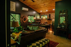 A cozy living room with a leather sofa, a dining area with a brick wall, and a kitchen with a hanging light fixture.