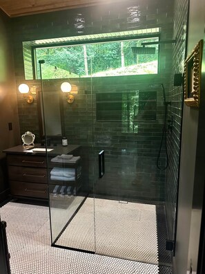 A modern bathroom with a glass shower enclosure, a sink with a mirror, and a window providing a view of greenery.
