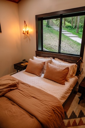 A bedroom with a bed, a nightstand, a wall-mounted light fixture, and a window with a view of a pathway and trees.