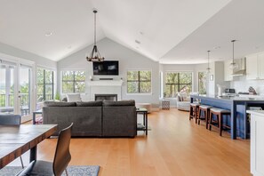 A spacious living area with a fireplace, a large sofa, a dining table, and a kitchen with a bar. - Cozy Lakefront Retreat. Family Friendly w/ Kayaks, Pool Access, Pickleball! (Spicewood)