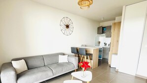 A modern living room with a grey sofa, a small white table with a vase of red flowers, and a kitchen in the background. - T1 bis - Quiet in La Rochelle (La Rochelle)