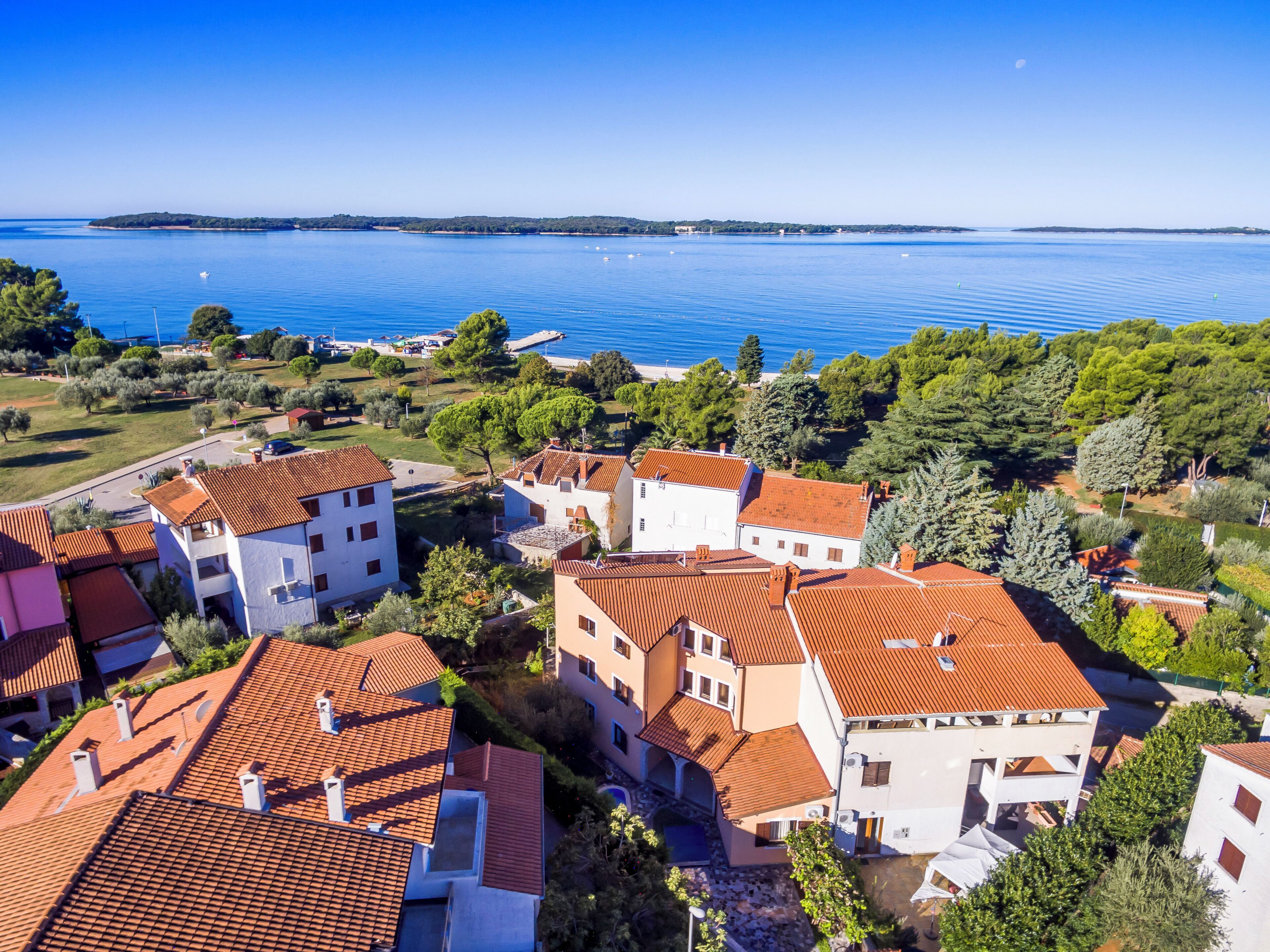 A coastal residential area with terracotta-roofed buildings, a marina, and a clear view of the sea.