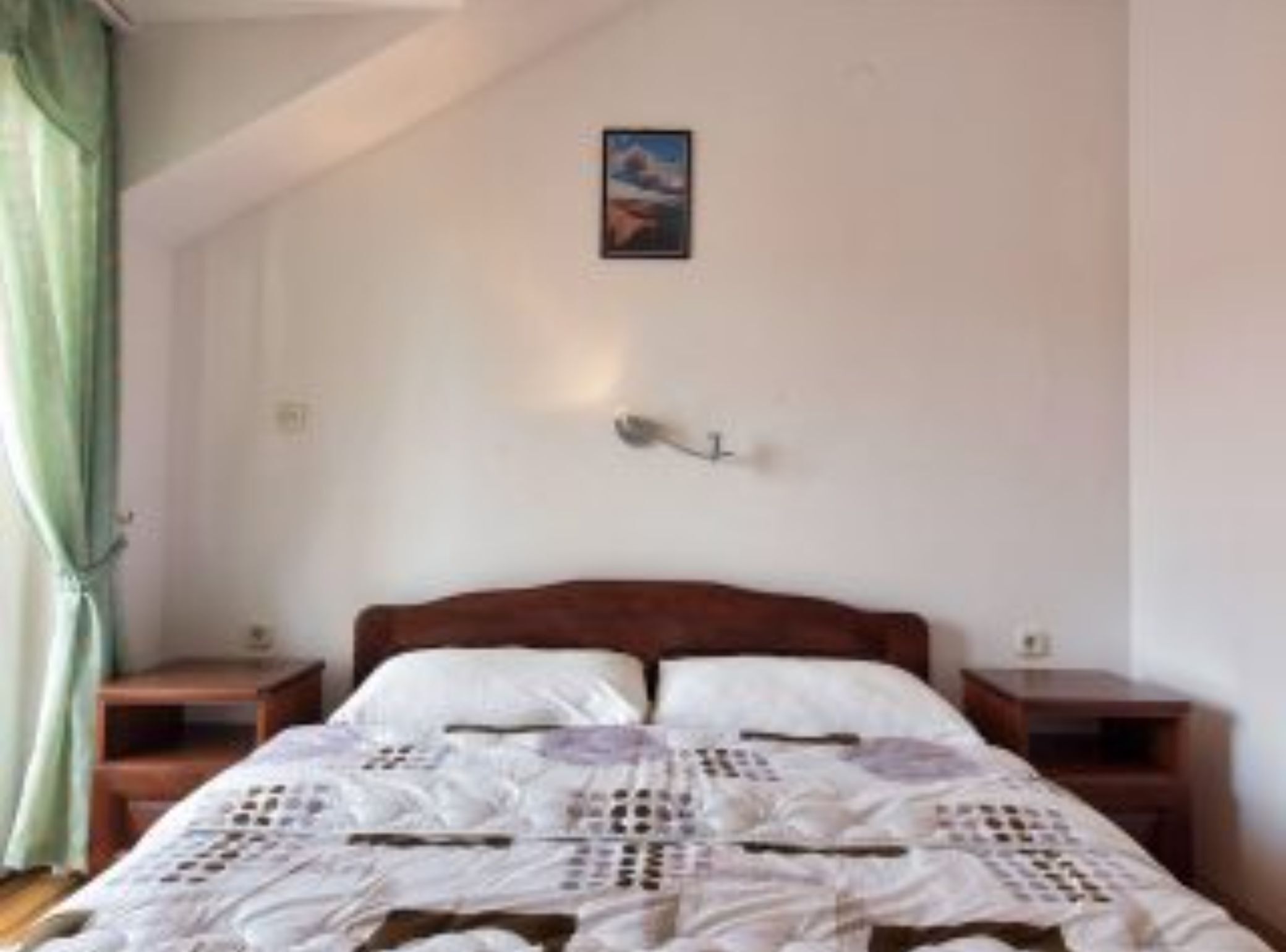 A bedroom with a wooden bed, a bedside table, a nightstand, a wall-mounted lamp, and a painting on the wall.