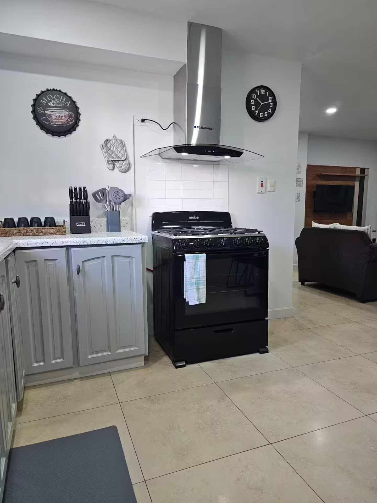 Una cucina con piano cottura, forno e zoccolo. Si vedono armadietti, una sveglia e un appendi-asciugamani a parete.