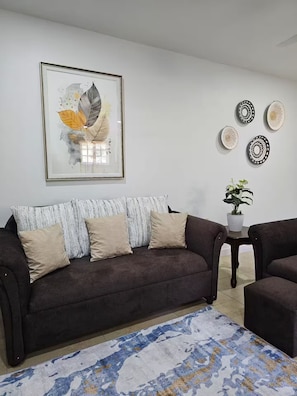 A living room with a brown sofa, a patterned rug, a framed artwork, and a small table with a plant.