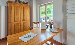 A wooden dining table with a newspaper, a chair, a cabinet with pottery, and a window with a view of a parking area.