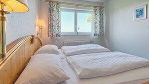 Quarto com cabeceira de madeira, duas camas brancas com almofadas, janela com cortinas florais, radiador e abajur à esquerda.
