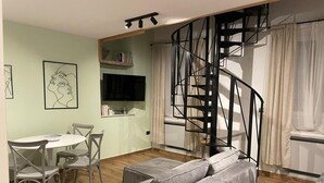 A living room with a grey sofa, a dining table with chairs, a TV, a bookshelf, and a spiral staircase. - Apartment Niccolo - Duplex Apartment with Sea View (Fazana)