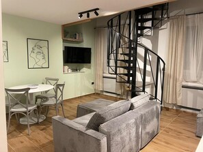 A living room with a grey sofa, a dining table with chairs, a TV, a bookshelf, and a spiral staircase. - Apartment Niccolo - Duplex Apartment with Sea View (Fazana)