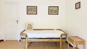 Ein Zimmer mit einem Einzelbett, einer Tür, zwei gerahmten Bildern an der Wand und einem kleinen Korbtisch.