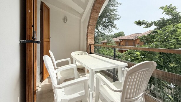 Ein Balkon mit weißem Tisch und Stühlen, einer Holztür und Blick auf Grünflächen und Gebäude.