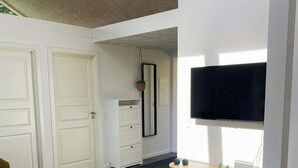 Ein Zimmer mit einem großen Flachbildfernseher an der Wand, einem Holztischchen mit einem Laptop und Pflanzen, einem Korbstuhl und einem weißen Kleiderschrank.