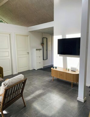 Et værelse med et stort fladskærms-tv på væggen, et trækonsolbord med en bærbar computer og planter, en fletstol og en hvid garderobe.