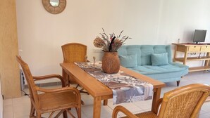 Uma sala de jantar com mesa de madeira, cadeiras de vime, um vaso trançado com plantas secas e um sofá azul claro ao fundo.
