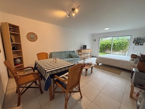 Ein modernes Wohnzimmer mit Esstisch, Stühlen, einer Couch, einem TV-Schrank und einem großen Fenster mit Blick auf Grünflächen.