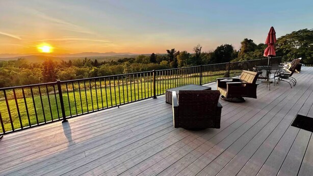 A wooden deck with outdoor furniture, a red umbrella, and a view of the sunset over a grassy landscape.