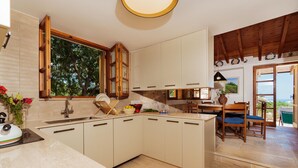 Een keuken met een groot raam, houten kastjes, een spoelbak en een eethoek met stoelen.