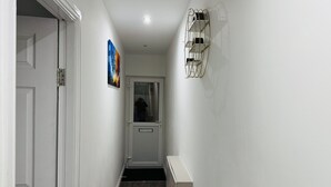 A narrow hallway with white walls, a wooden floor, a door at the end, and a small shelf with items on the right wall. - Modern 1BR Home in Clayton-le-Moors (Clayton Le Moors)