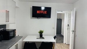 Eine moderne Küche mit einem Flachbildfernseher, der 'NETFLIX' anzeigt, ein Essbereich mit Tisch und Stühlen sowie ein Flur, der in einen anderen Raum führt.