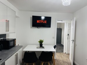 A modern kitchen with a flat-screen TV displaying 'NETFLIX,' a dining area with a table and chairs, and a hallway leading to another room. - Modern 1BR Home in Clayton-le-Moors (Clayton Le Moors)