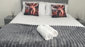 Ein ordentlich bezogenes Bett mit einer grauen, strukturierten Tagesdecke und zwei mit Kuhmuster versehenen Kissen, ein weißer Nachttisch mit Lampe sowie ein an der Wand hängendes Bild.