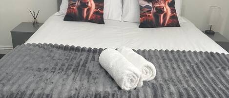 Ein ordentlich bezogenes Bett mit einer grauen, strukturierten Tagesdecke und zwei mit Kuhmuster versehenen Kissen, ein weißer Nachttisch mit Lampe sowie ein an der Wand hängendes Bild.