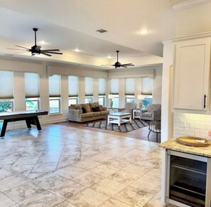 A spacious living area with a pool table, a sofa, and a chair. There are ceiling fans and large windows.