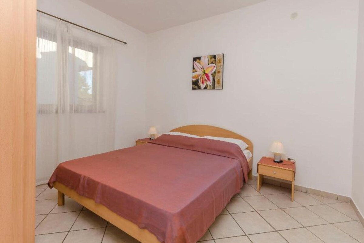 A bedroom with a bed, two bedside tables, a window with curtains, and a wall-mounted picture.