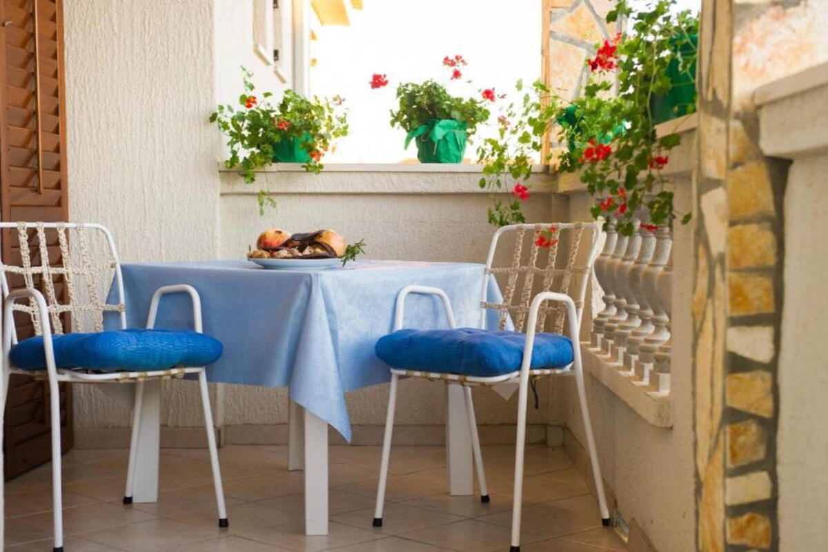 A table set for two with a blue tablecloth, two white chairs with blue cushions, and a window with potted plants and flowers.