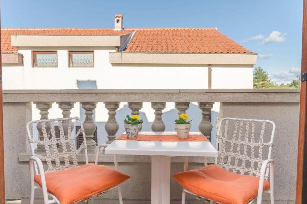 A balcony with two wicker chairs and a small table, a potted plant, and a building with a tiled roof in the background.