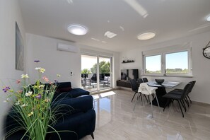 Ein modernes Wohnzimmer mit einer Couch, einem Esstisch mit Stühlen, einem großen Fenster und einer Deckenleuchte.