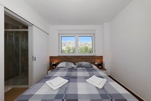 A bedroom with a bed, two bedside tables, a window with a mountain view, and a glass shower enclosure. - Apartment Lux 1 near the Sea with Balcony (Starigrad)