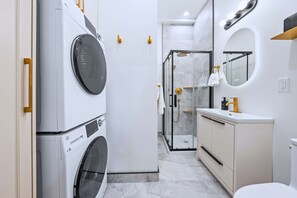 Kompakti ja moderni kylpyhuone, jossa on pyykinpesukone, lasinen suihkuseinä, peilillä varustettu allaskaappi ja wc-istuin.