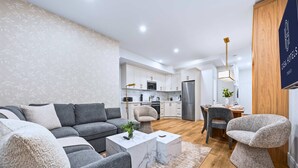 Ein modernes Wohnzimmer mit einem grauen Ecksofa, einem Marmortisch und einem außergewöhnlichen Sessel. Im Hintergrund ist eine Küche mit weißen Schränken und Edelstahlgeräten zu sehen.