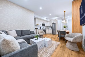 Un salon moderne avec un canapé d’angle gris, une table basse en marbre et un fauteuil original. On aperçoit à l’arrière-plan une cuisine avec des meubles blancs et des appareils électroménagers en acier inoxydable.