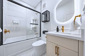 Une salle de bain moderne avec une douche à parois de verre, des toilettes blanches, un meuble-lavabo avec un robinet doré et un miroir rond.