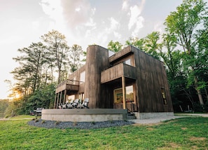 A modern wooden house with a balcony, outdoor seating, and a view of the surrounding forest.