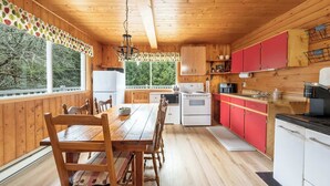 A wooden cabin interior with a dining table, chairs, a kitchen with red cabinets, and a view through large windows.