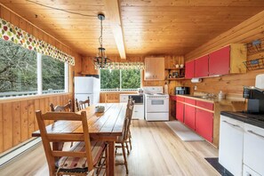 A wooden cabin interior with a dining table, chairs, a kitchen with red cabinets, and a view through large windows. - Spa, Nature & Adventure | Chalet Le Vagabond (Saint-Léon-De-Standon)