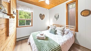 A bedroom with a wooden ceiling, a bed with a green blanket, a ceiling fan, and a window with a view of greenery.