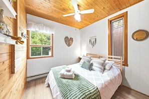 A bedroom with a wooden ceiling, a bed with a green blanket, a ceiling fan, and a window with a view of greenery. - Spa, Nature & Adventure | Chalet Le Vagabond (Saint-Léon-De-Standon)