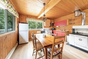 A wooden cabin kitchen with a wooden table, chairs, a refrigerator, a stove, and a window overlooking greenery. - Spa, Nature & Adventure | Chalet Le Vagabond (Saint-Léon-De-Standon)